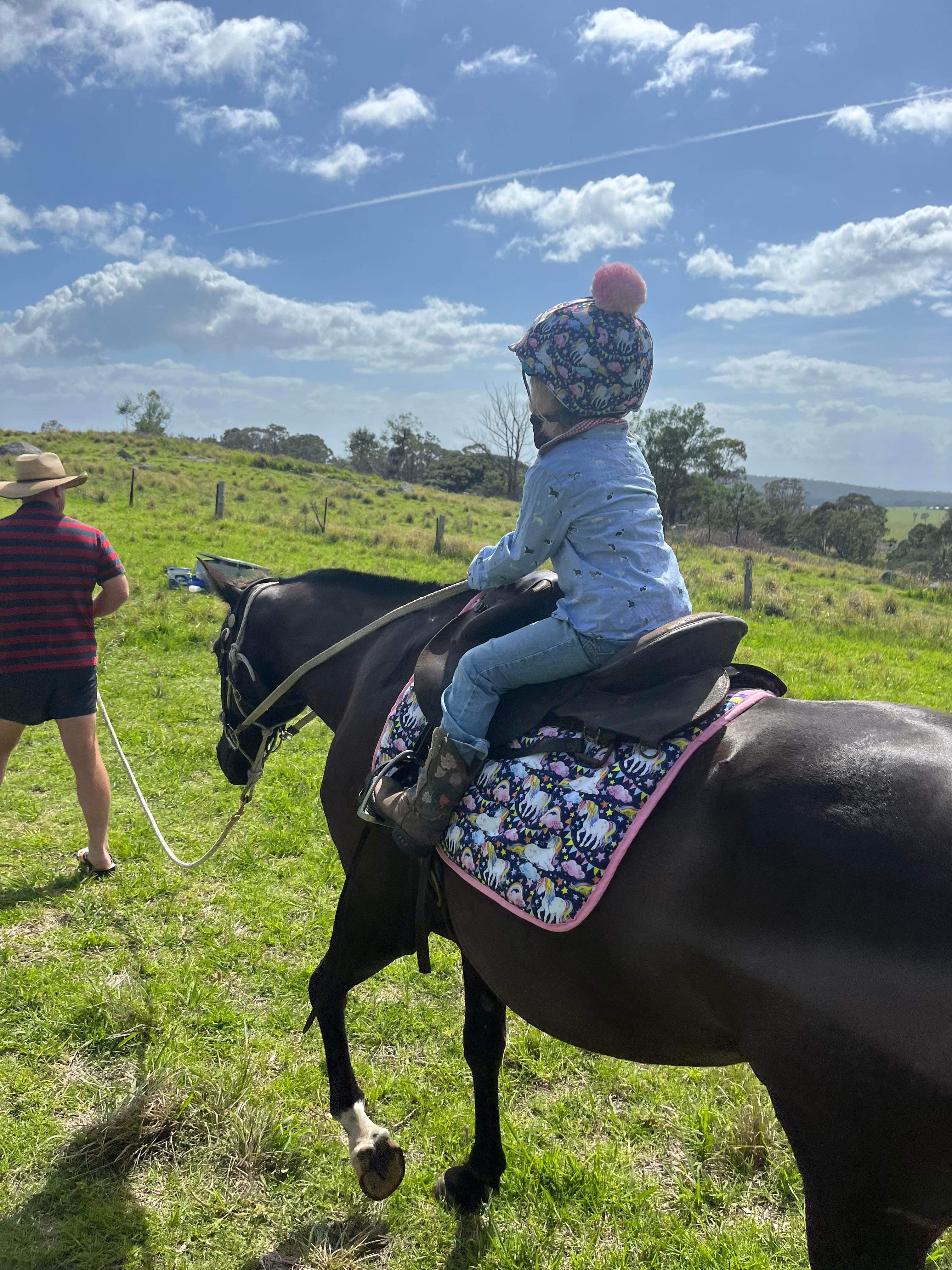 A young equestrian riding a black horse in a green paddock wearing a unicorn helmet cover with a pink pom pom and a matching saddle pad